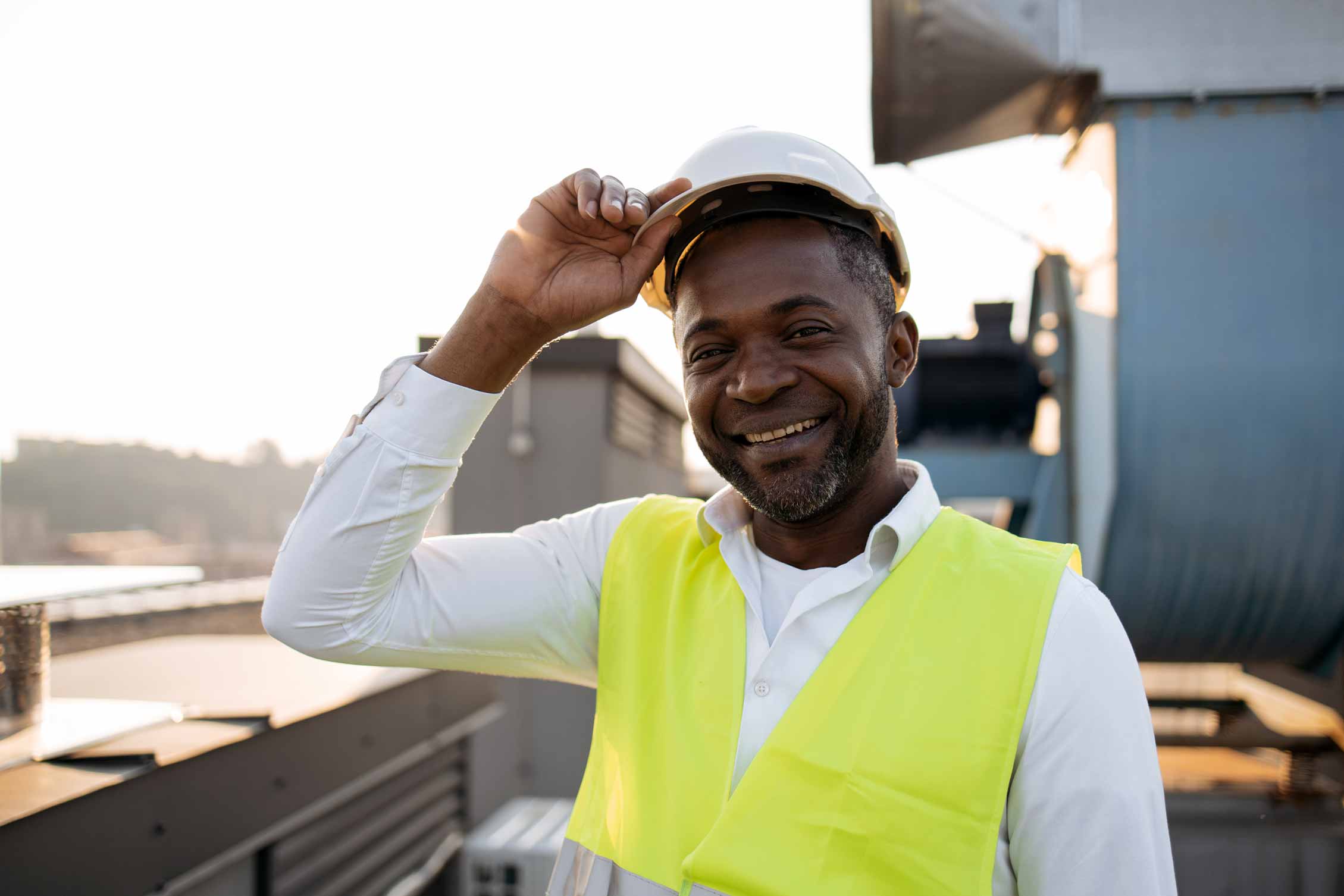 A man wearing a white shirt, reflective vest, and hard hat, smiling confidently on a worksite.