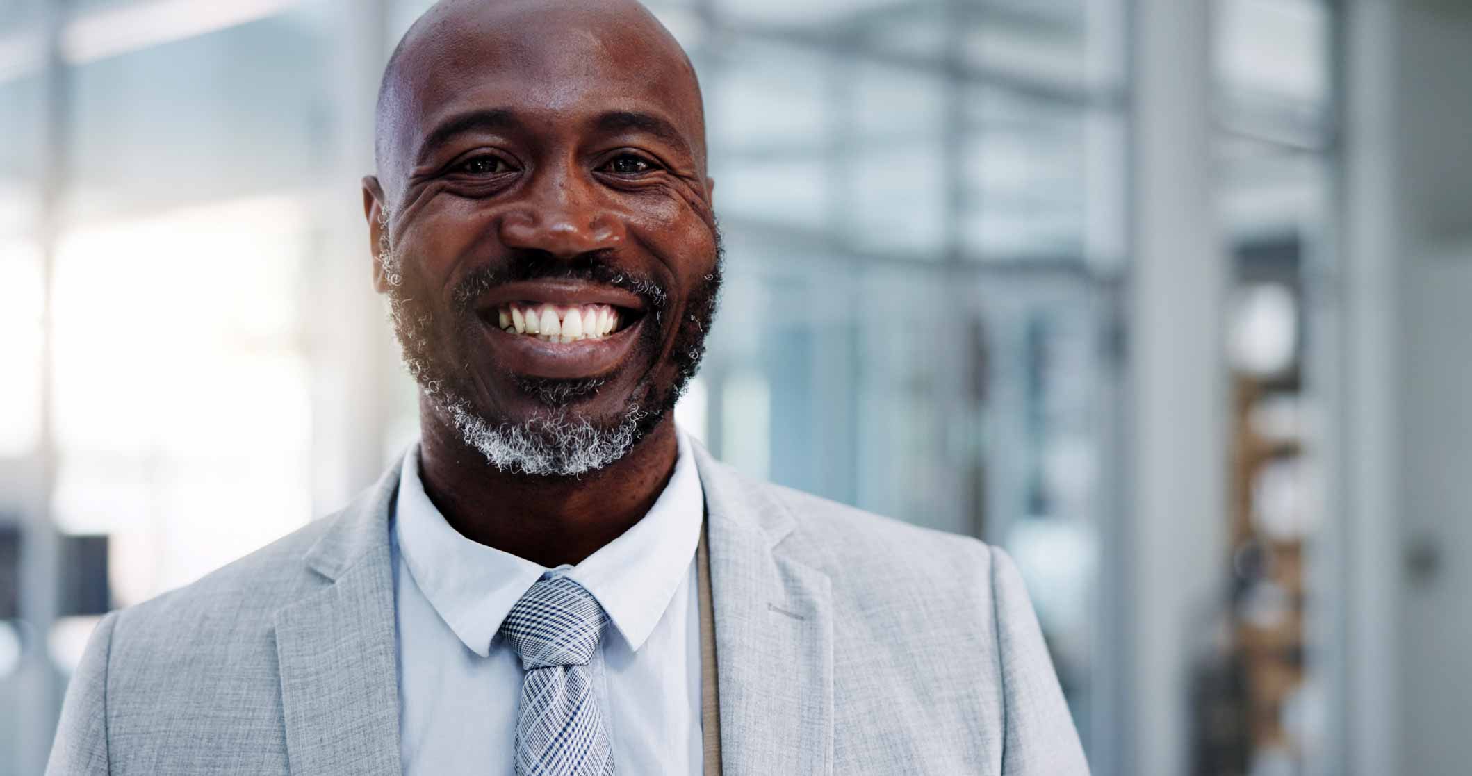 A man wearing a light gray suit and tie, smiling in a bright office environment.