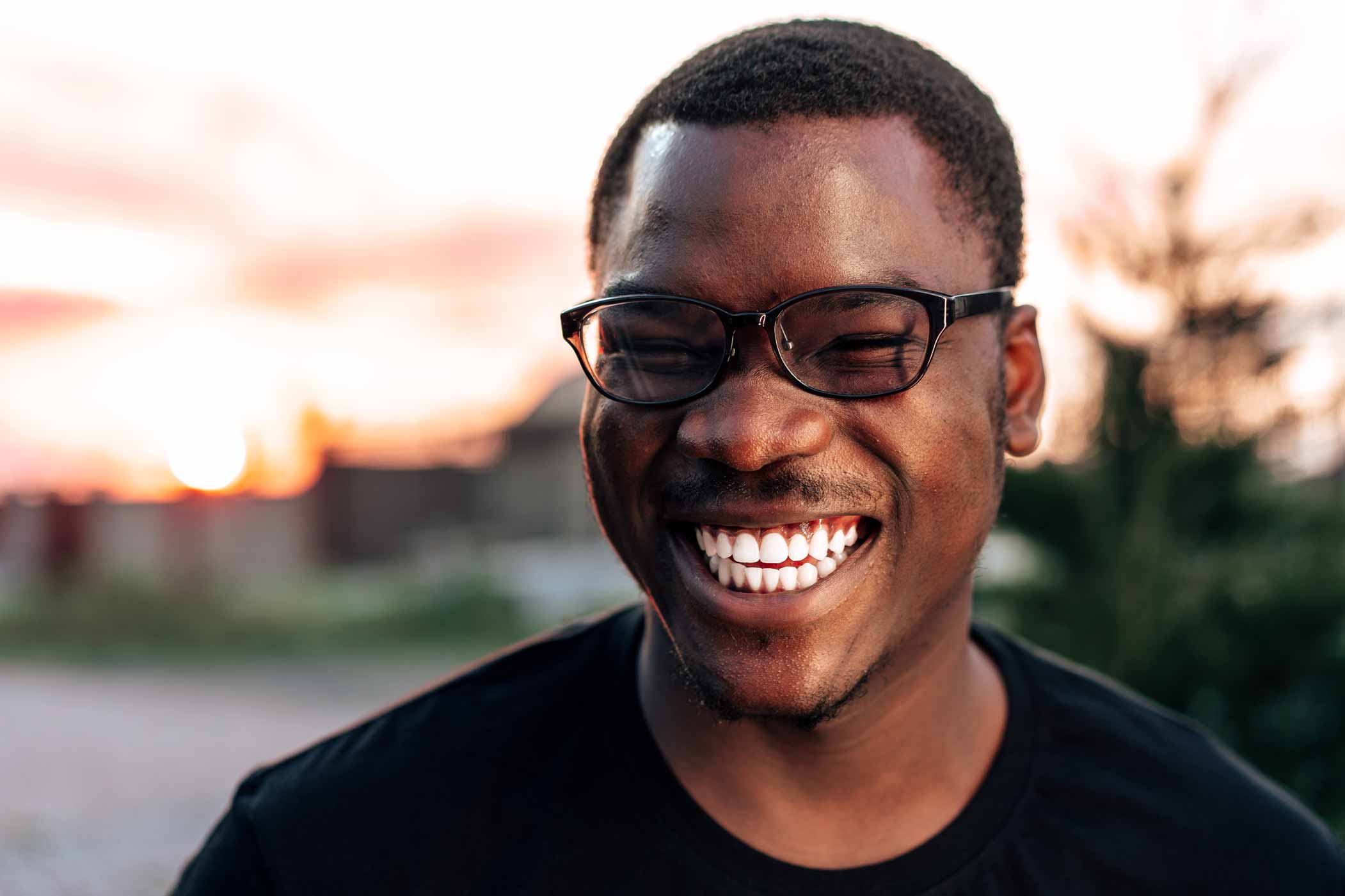 A young man wearing glasses, laughing with a sunset in the background.